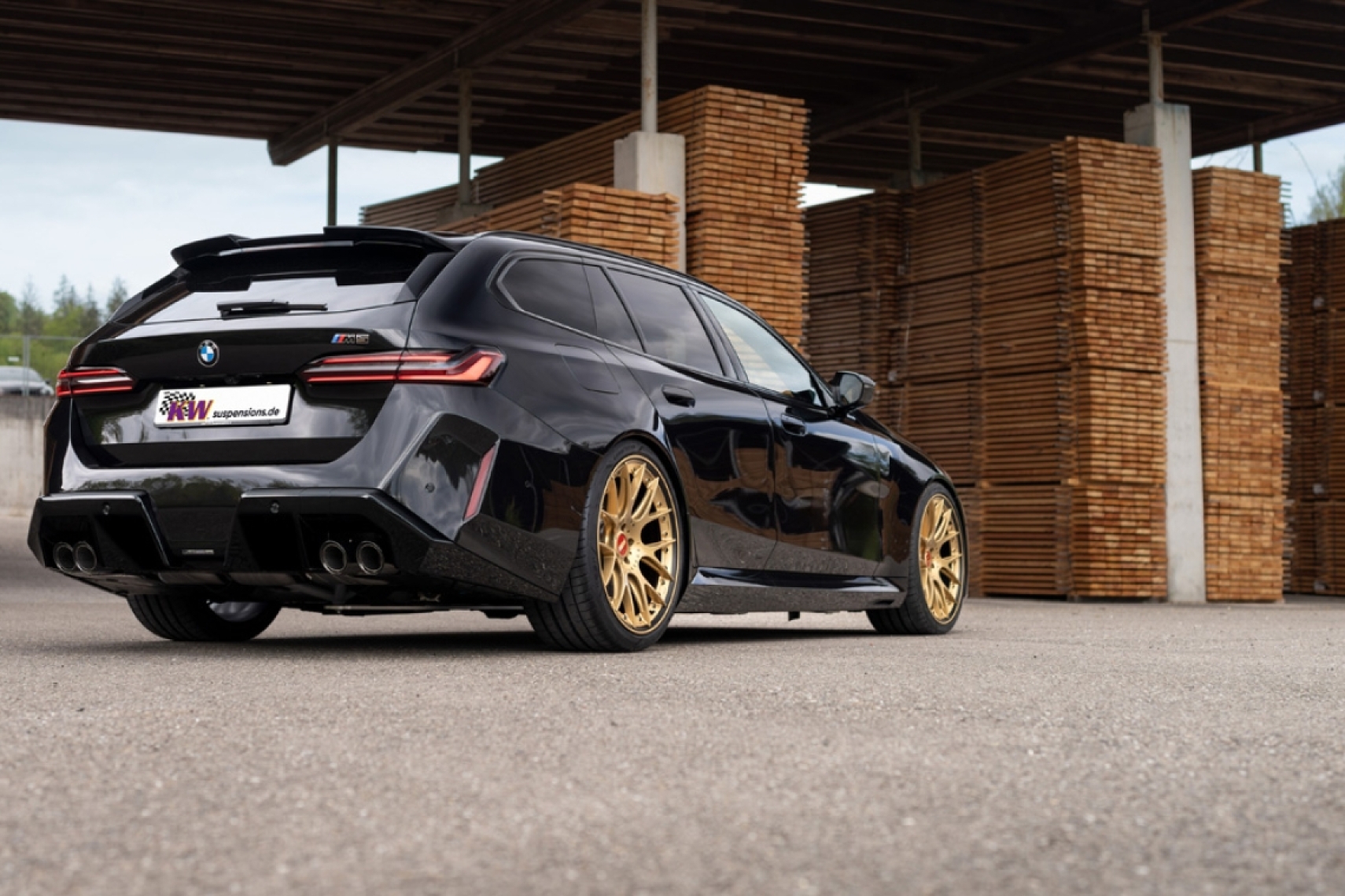 BMW M5 Touring G99 lowered on KW height-adjustable springs with gold wheels, shown from the rear three-quarter angle.